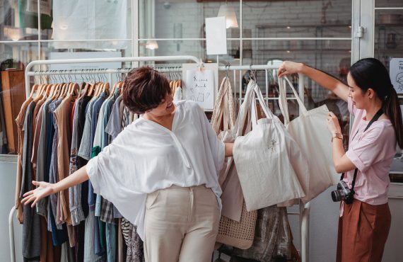 Young content ethnic female buyers selecting fabric bags on rack in local urban shop in daylight