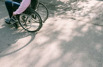 A person in a wheelchair navigating a sunlit road with tree shadows. The image captures a moment of mobility and independence.