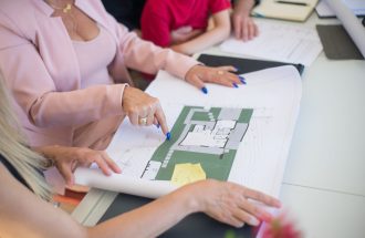 A real estate agent in a pink blazer shows floor plans to clients, discussing property details.