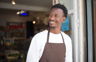 Smiling store owner in brown apron standing at entrance, welcoming customers.