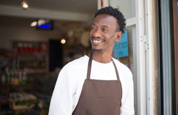 Smiling store owner in brown apron standing at entrance, welcoming customers.