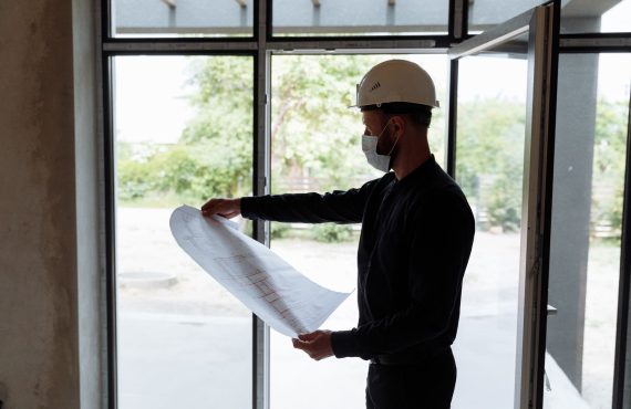 Architect reviewing floor plans indoors, wearing safety gear, focused on construction planning.