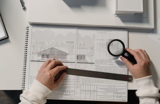 Hands holding a ruler and magnifying glass over architectural floor plans on a white table.