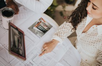 A woman reviews architectural floor plans on a table with a tablet in a modern workspace.
