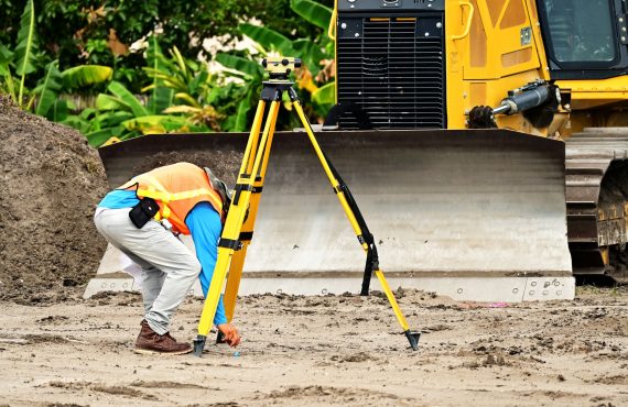 A construction worker operates a total station in front of a bulldozer on a construction site.