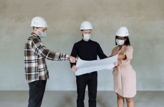 Three architects wear masks and helmets while reviewing construction plans indoors.
