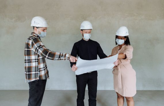 Three architects wear masks and helmets while reviewing construction plans indoors.