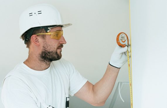 Construction worker with hard hat and goggles measuring a wall with a tape measure.