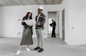 Two architects examine blueprints in a partially finished building while discussing design plans.